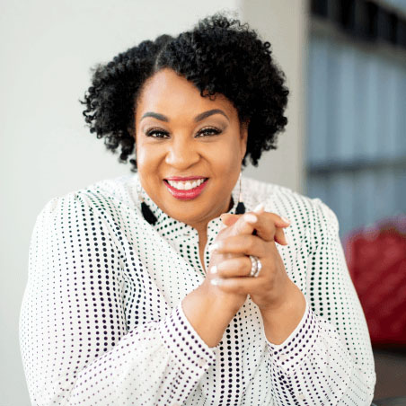 Photo of Tamika, a middle-aged black woman with short curly hair. She is wearing a white striped shirt and smiling with her hands clasped.