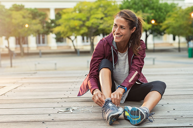 Photo of a young white woman with brown hair in a ponytail wearing athletic clothing. She is sitting outside on a boardwalk in front of a row of trees and is tying one of her running shoes.