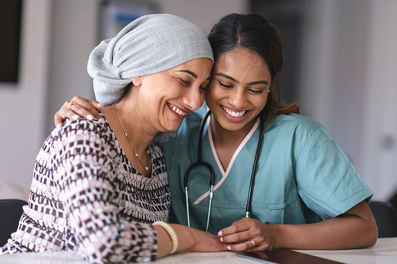 Photo of two women indoors, one older wearing a gray headwrap and patterned shirt and the other wearing scrubs and a stethoscope. They are close and smiling together while looking down.