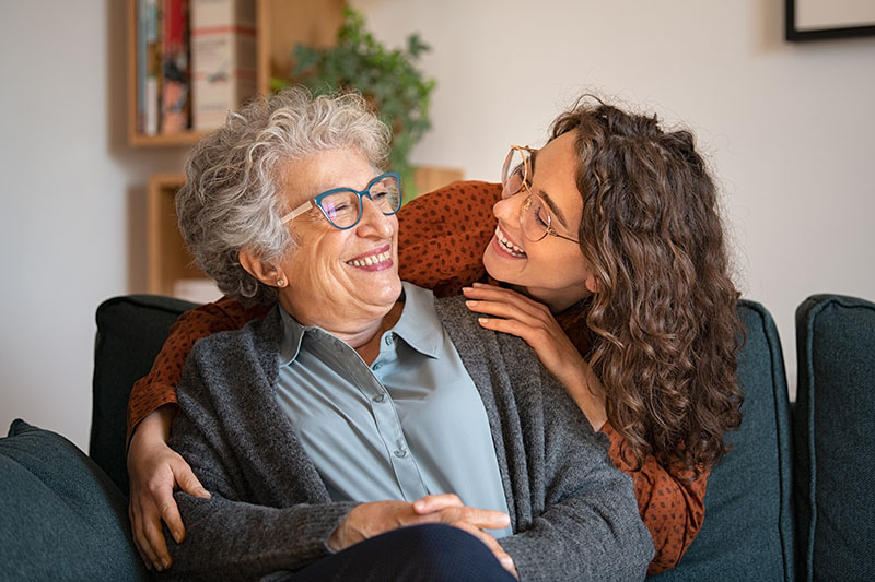 Photo of an older woman with gray hair wearing a gray sweater and shirt. A young woman with brown hair and a red shirt is hugging the older woman from behind and smiling at her. Both women are wearing glasses.