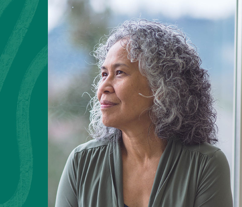 Image of an older woman with medium skin and gray curly hair to her shoulders. She is wearing a gray top and is smiling softly while looking to the left.