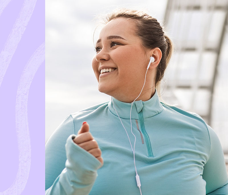 Image of a younger white woman with brown hair in a ponytail, wearing a light blue quarter-zip top and white earbuds. She is jogging and smiling while looking to the left.