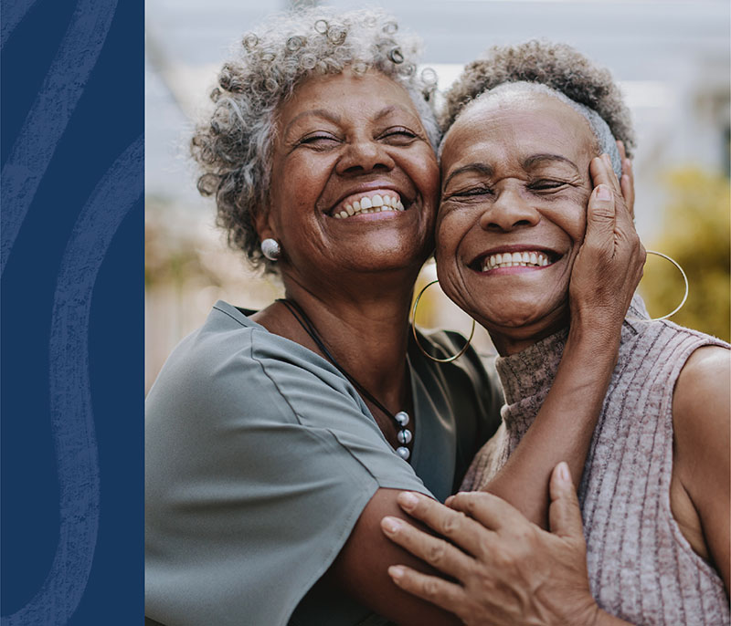 Image of two older black women with gray curly hair, smiling and embracing each other. One is wearing a green t-shirt, while the other has a light gray tank top.