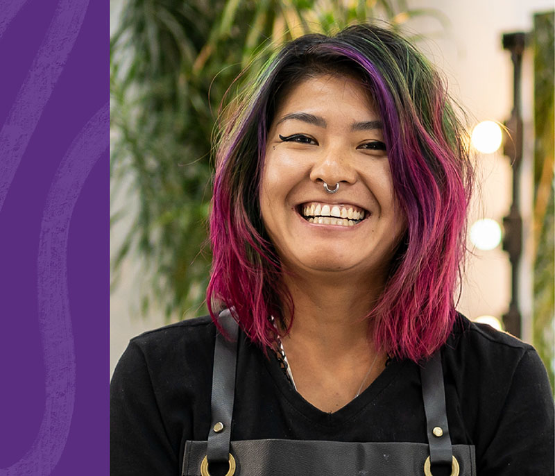 Image of a younger Asian woman with shoulder-length black and pink hair, wearing a black top and apron. She has a nose piercing and is smiling at the camera.