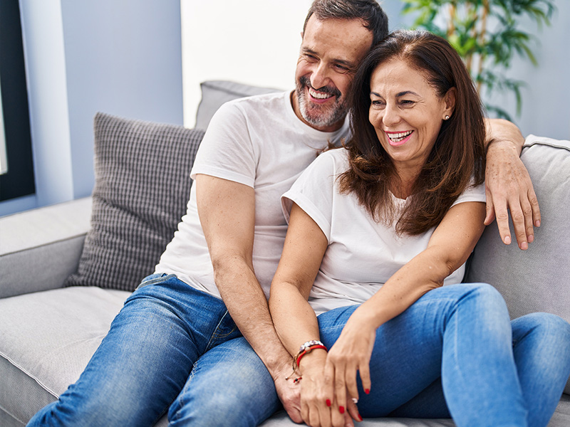 A smiling couple is sitting close together on a couch, holding hands and leaning into each other, both dressed in white t-shirts and jeans. 