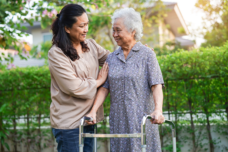 Photo of two women standing outside in front of a row of green bushes. On the left is a younger woman with black hair in a ponytail wearing a tan shirt. She is facing and holding the arm of an older woman with short gray hair wearing a long dress and holding a walker. They are both smiling at each other.