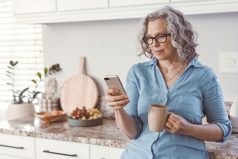 Photo of an older woman with gray curly hair leaning against a marble kitchen counter. She is smiling softly looking at her mobile phone in one hand with a mug in the other.