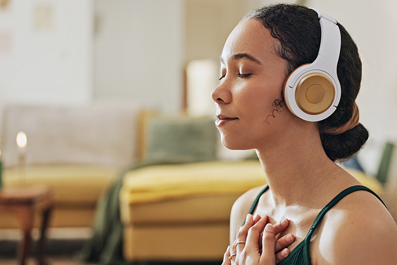 Photo of a young woman with a medium skin tone and black hair in a bun, sitting in a room in front of a yellow couch. She is wearing white headphones and has her eyes closed, holding her hands on her chest.