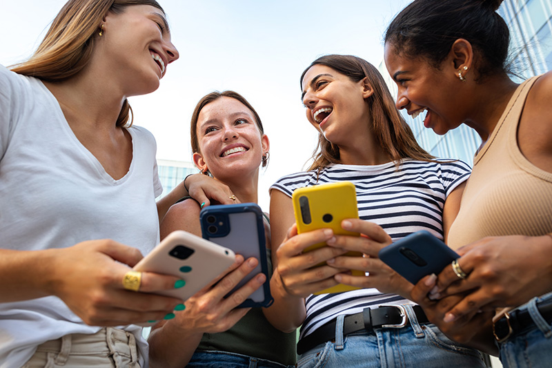 Photo of a group of four young women outside from a low angle. They are laughing together and holding mobile phones.
