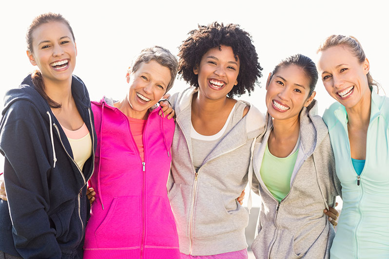 Photo of five women standing with their arms around each other and smiling, wearing athletic clothing. The women range in age from early 20s to 60s and have different skin tones.