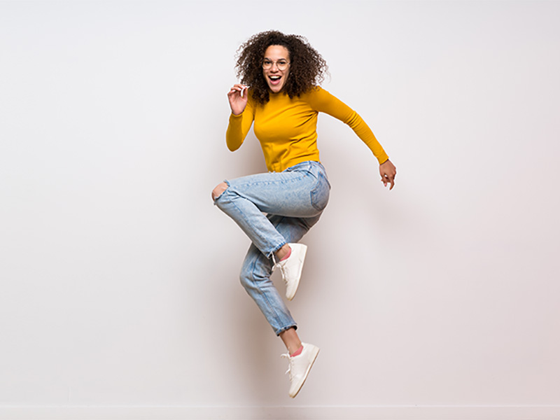 A person with curly hair and glasses jumps joyfully in a yellow shirt and ripped jeans against a white background.