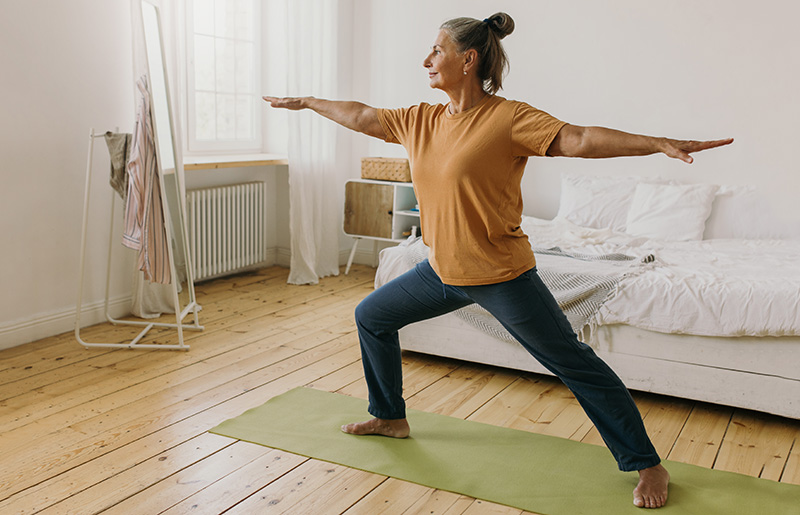 Person practicing yoga in a bedroom, standing on a green mat in a warrior pose.