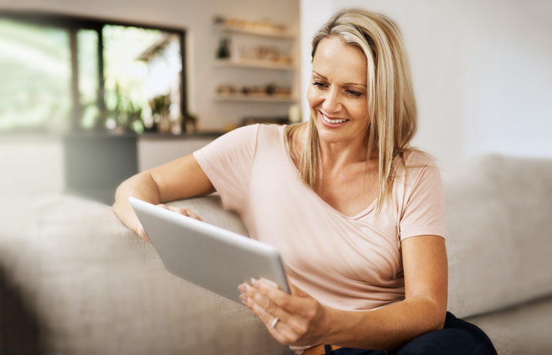 A woman smiling while looking at a tablet in a bright, cozy living room.