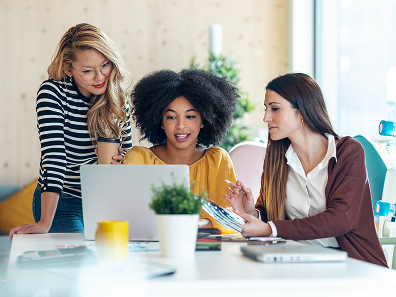 Three women collaborate at a table with a laptop, coffee, and documents.