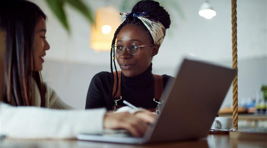 Mujer con una camisa negra sentada y mirando a una joven con una camisa blanca, que está usando una computadora portátil.