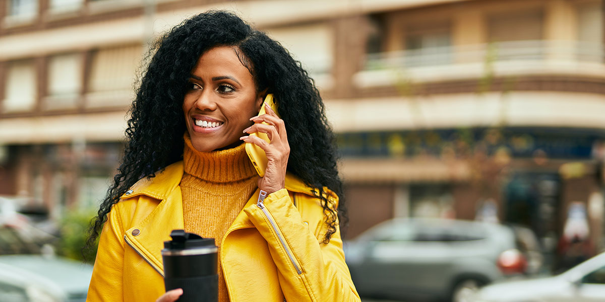 A woman wearing a bright yellow jacket and holding a travel mug is smiling as she talks on her phone while walking. 