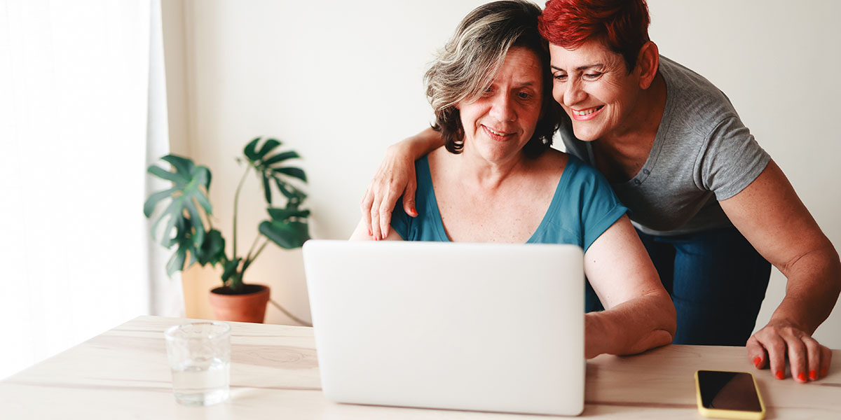 Two women are smiling and looking at a laptop screen together, with one standing and embracing the other from behind.