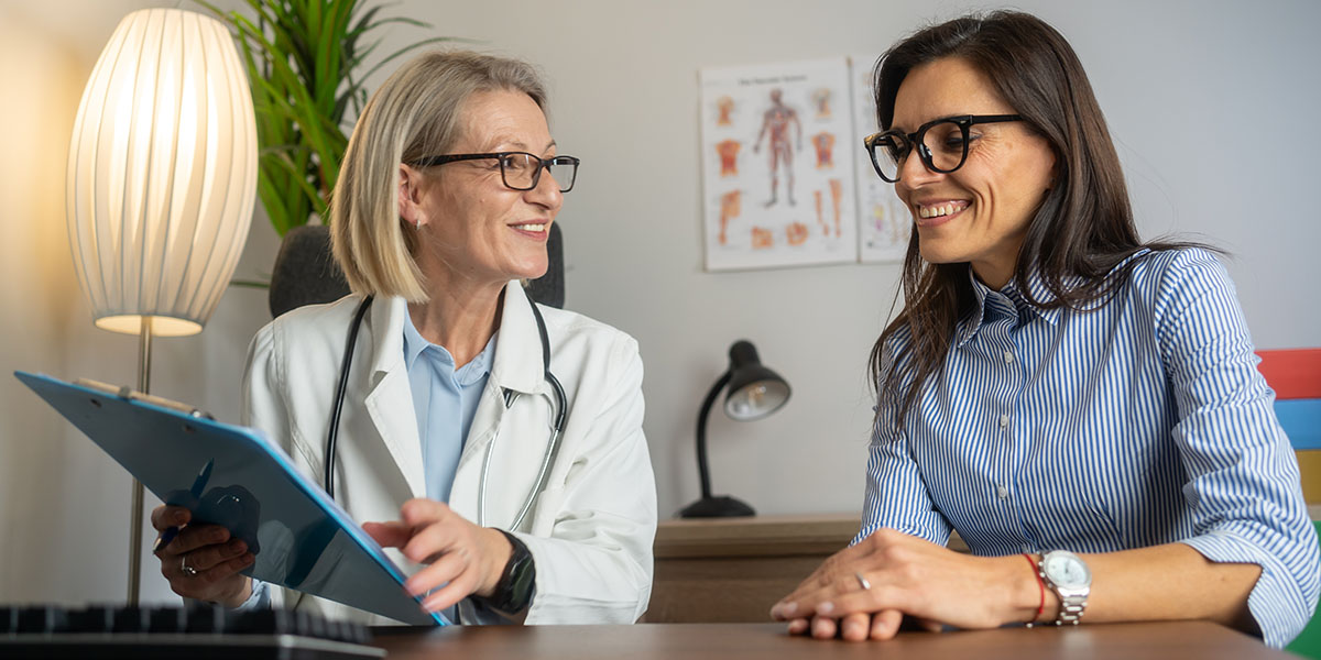 A doctor and a patient are sitting at a desk, both smiling and engaged in a conversation, with the doctor holding a clipboard and the patient looking at the clipboard.