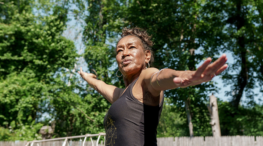 A woman stands outdoors with arms outstretched, surrounded by green trees.