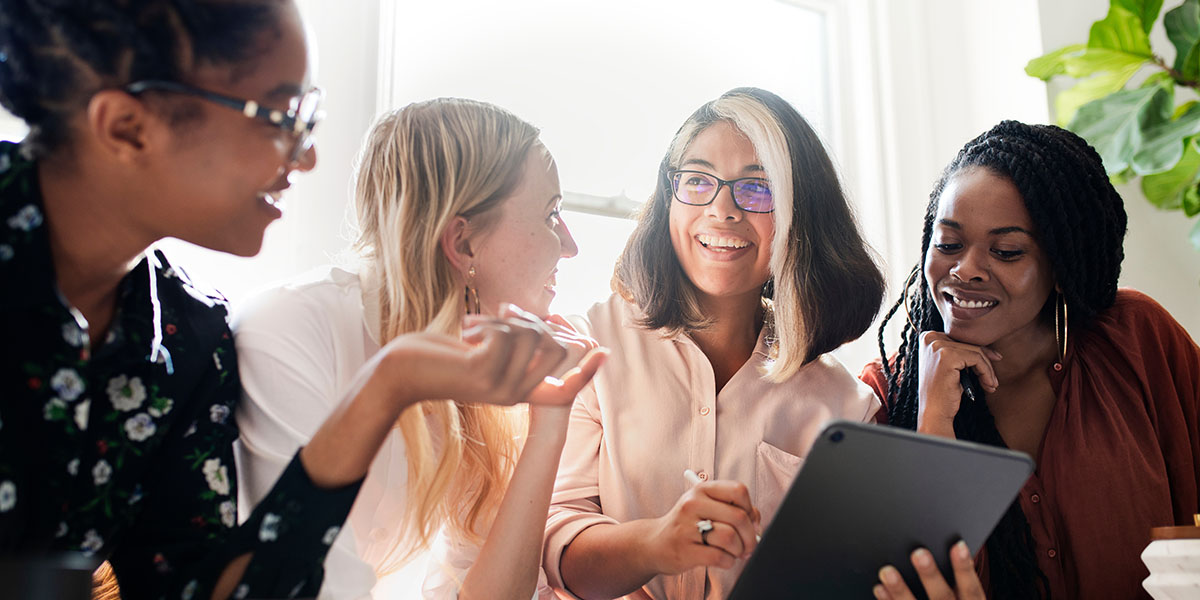 Un grupo diverso de cuatro mujeres está reunido alrededor de una mesa, sonriendo y participando en una charla, mientras una mujer sostiene una tableta.