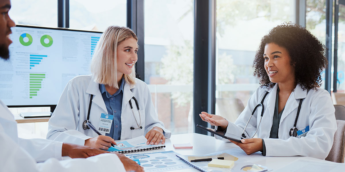 Un grupo de profesionales de la salud con batas de laboratorio hablan alrededor de una mesa llena de documentos y gráficos.