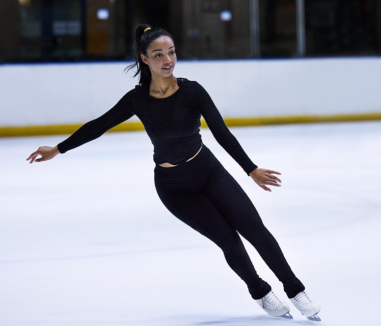 A young female figure skater in a black outfit glides across an ice rink. She has a focused expression, her arms extended, and her hair pulled back in a ponytail. The background is slightly blurred, showing the rink's boards and lights.