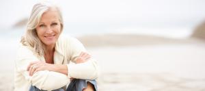 Smiling white-haired woman sitting on a beach with her arms folded over her knees.