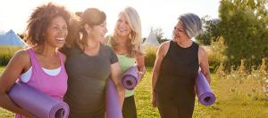 Four women walk through a meadow carrying rolled purple yoga mats and laughing together.